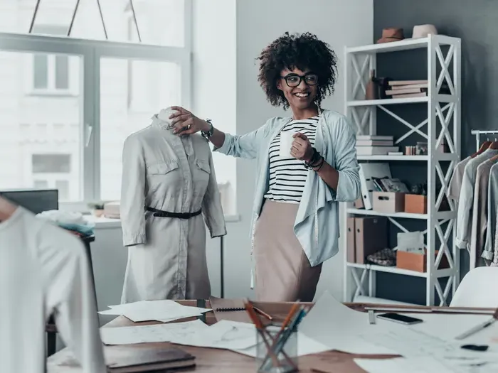 A smiling clothing designer standing beside a mannequin in her fashion studio surrounded by sketches and fabric samples